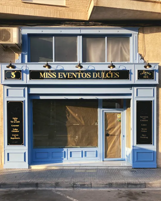 Fachada de Miss Eventos Dulces en azul claro, con molduras, rótulos negros con letras doradas, escaparate y puerta acristalada.
